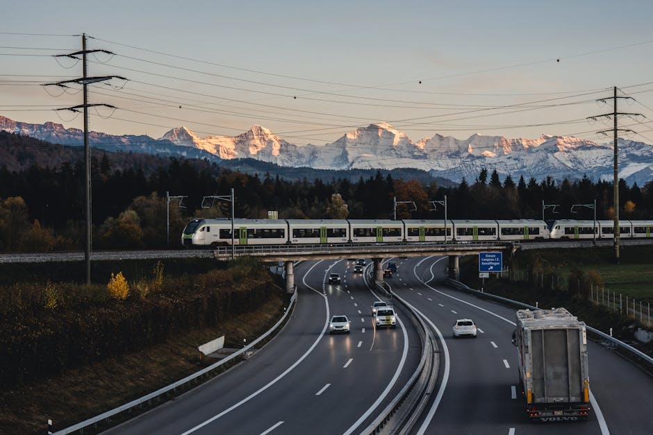 A train crosses a bridge over a highway near Bern, Switzerland at sunset with mountains in the background.